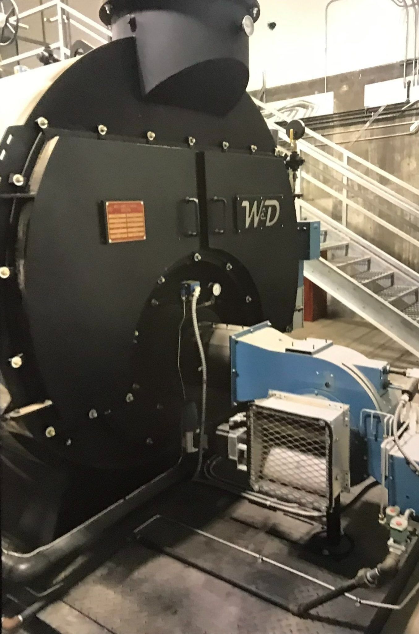 Large black boiler with a blue burner, next to stairs in an industrial setting.
