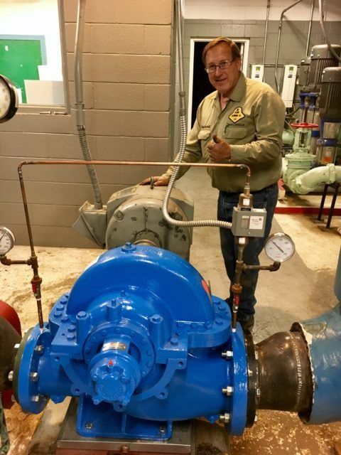 Man in work clothes gives a thumbs up next to a large blue water pump in an industrial setting.