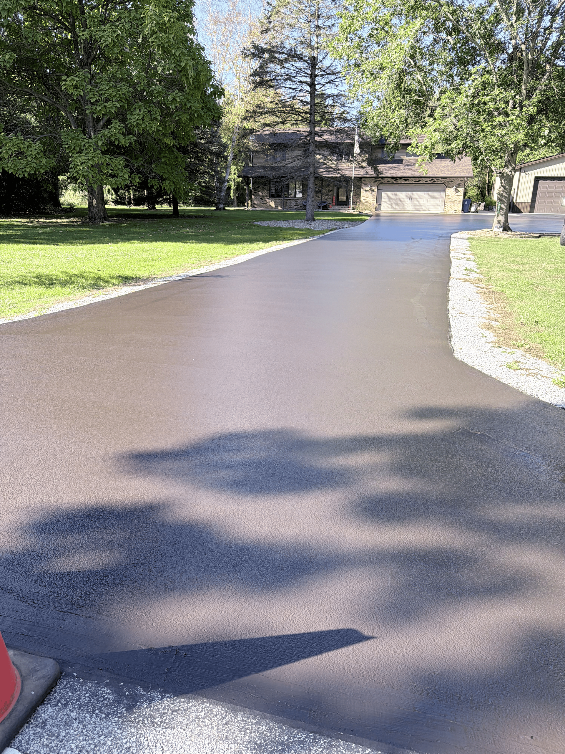 Freshly paved asphalt driveway leading towards a two-story house, bordered by grass and trees.