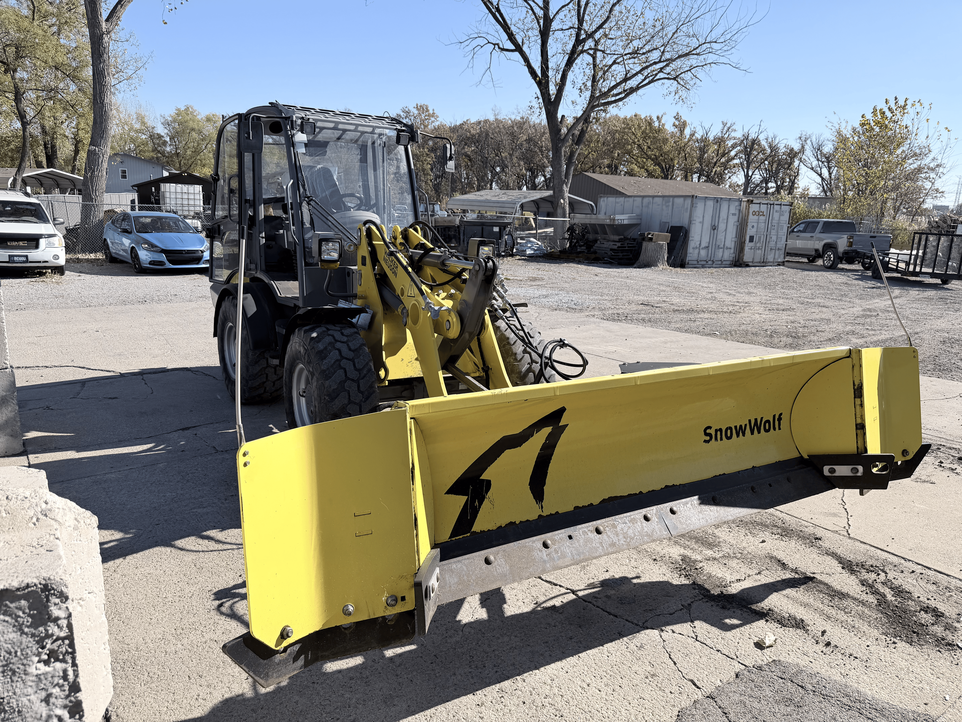 Yellow snowplow attached to a black tractor on a paved lot on a sunny day.