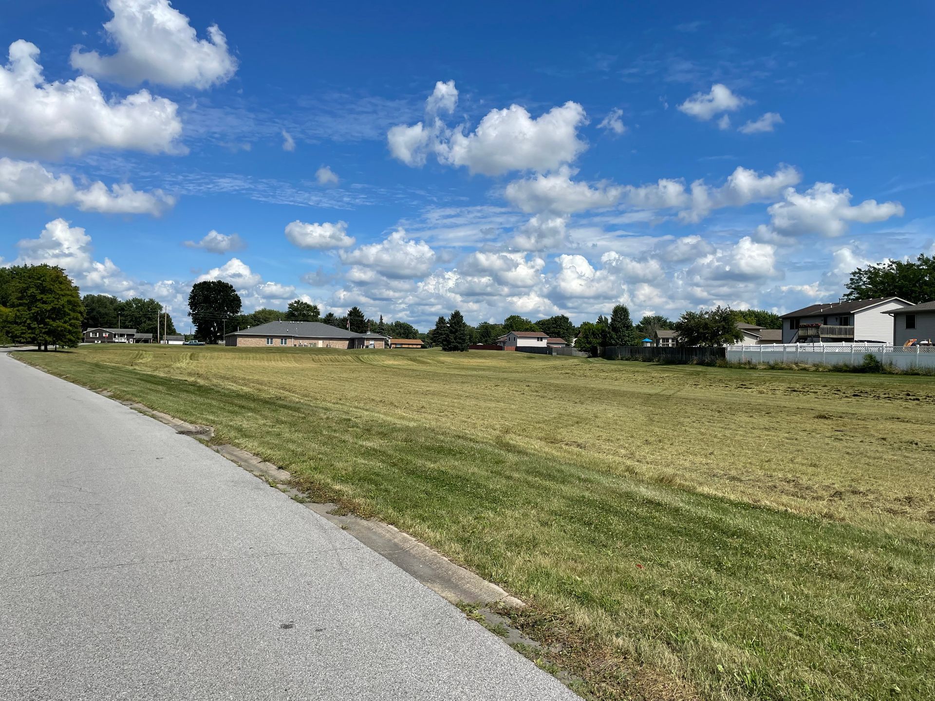 Open grassy lot with sparse vegetation under a blue sky dotted with clouds, bordered by a road.