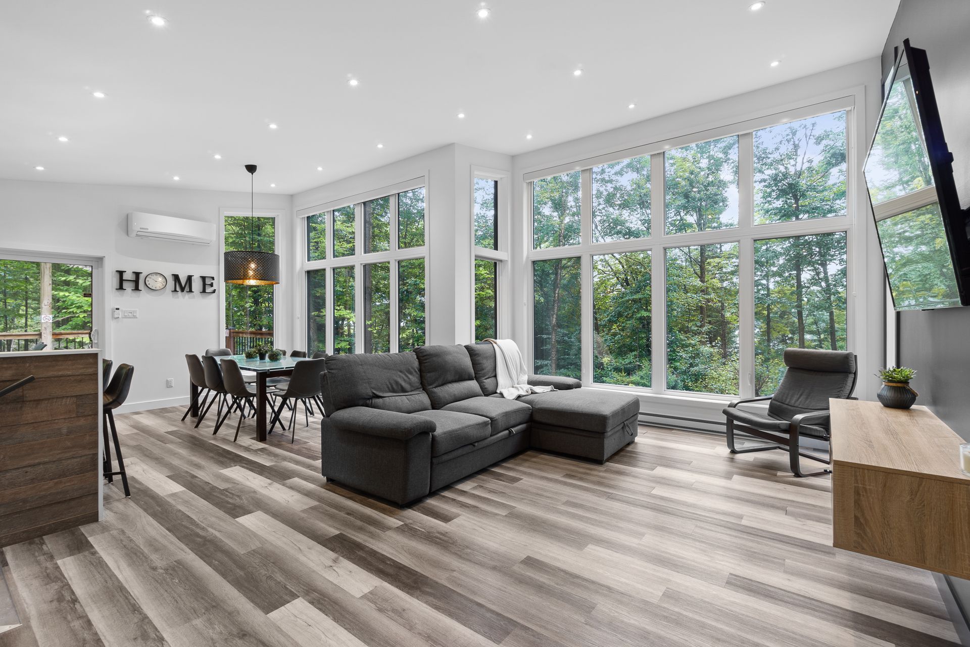 Salon spacieux avec de grandes fenêtres, canapé gris foncé, table à manger et parquet.