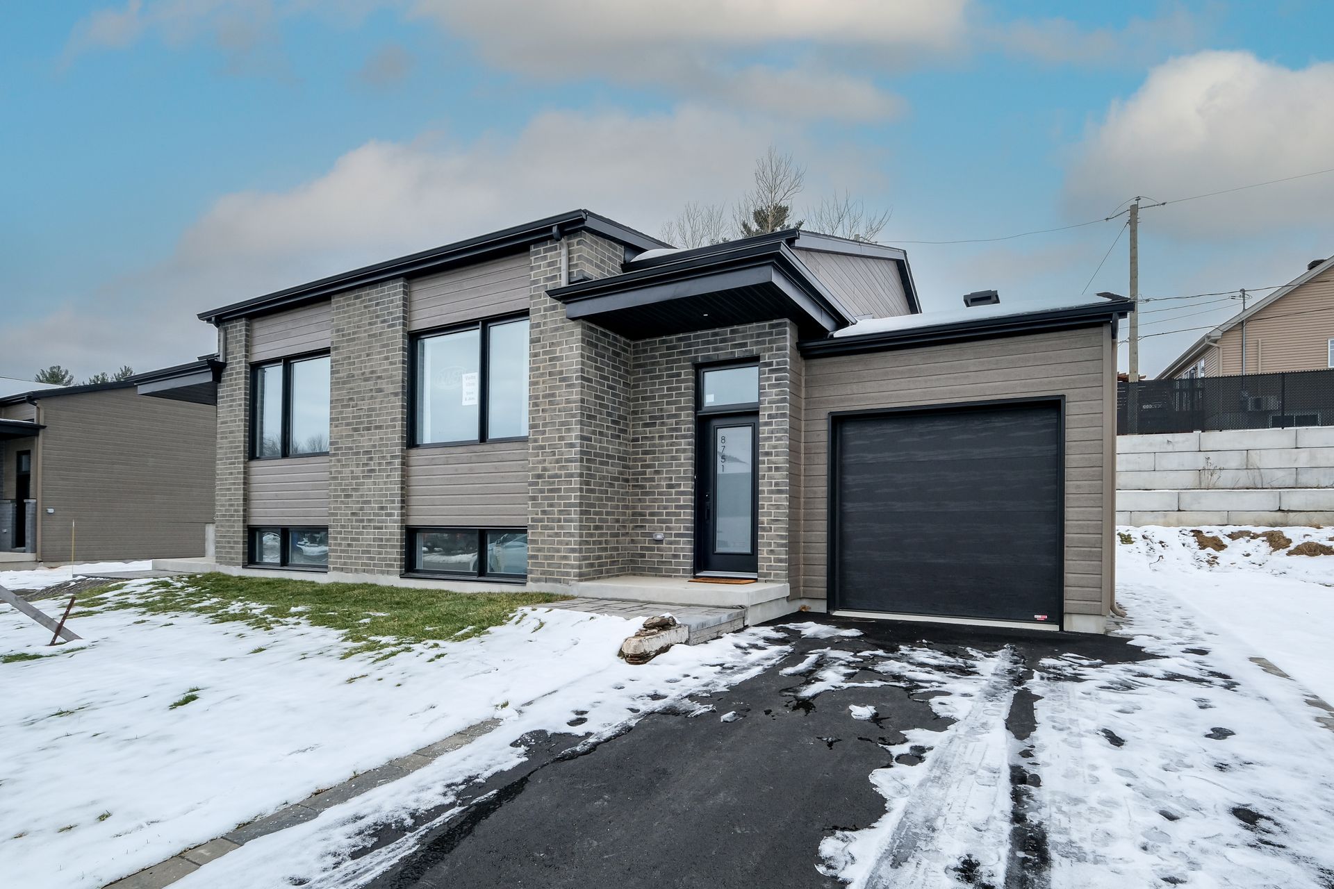 Maison moderne avec revêtement en brique et en bois, cour enneigée et porte de garage noire.