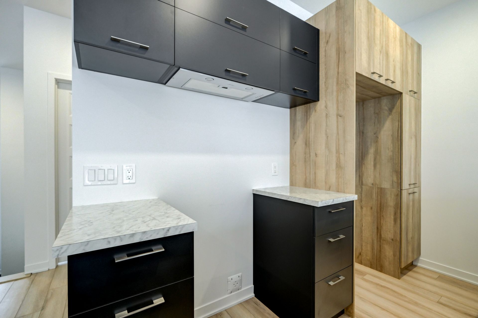 Cuisine moderne avec armoires noires et bois, comptoir blanc, planchers de bois clair.