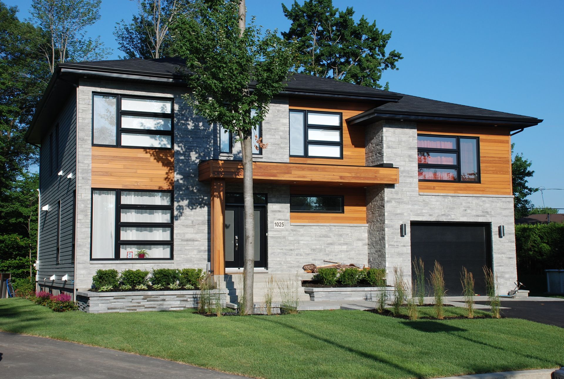 Maison moderne à deux étages avec pierre grise, accents de bois, fenêtres noires et porte de garage noire.