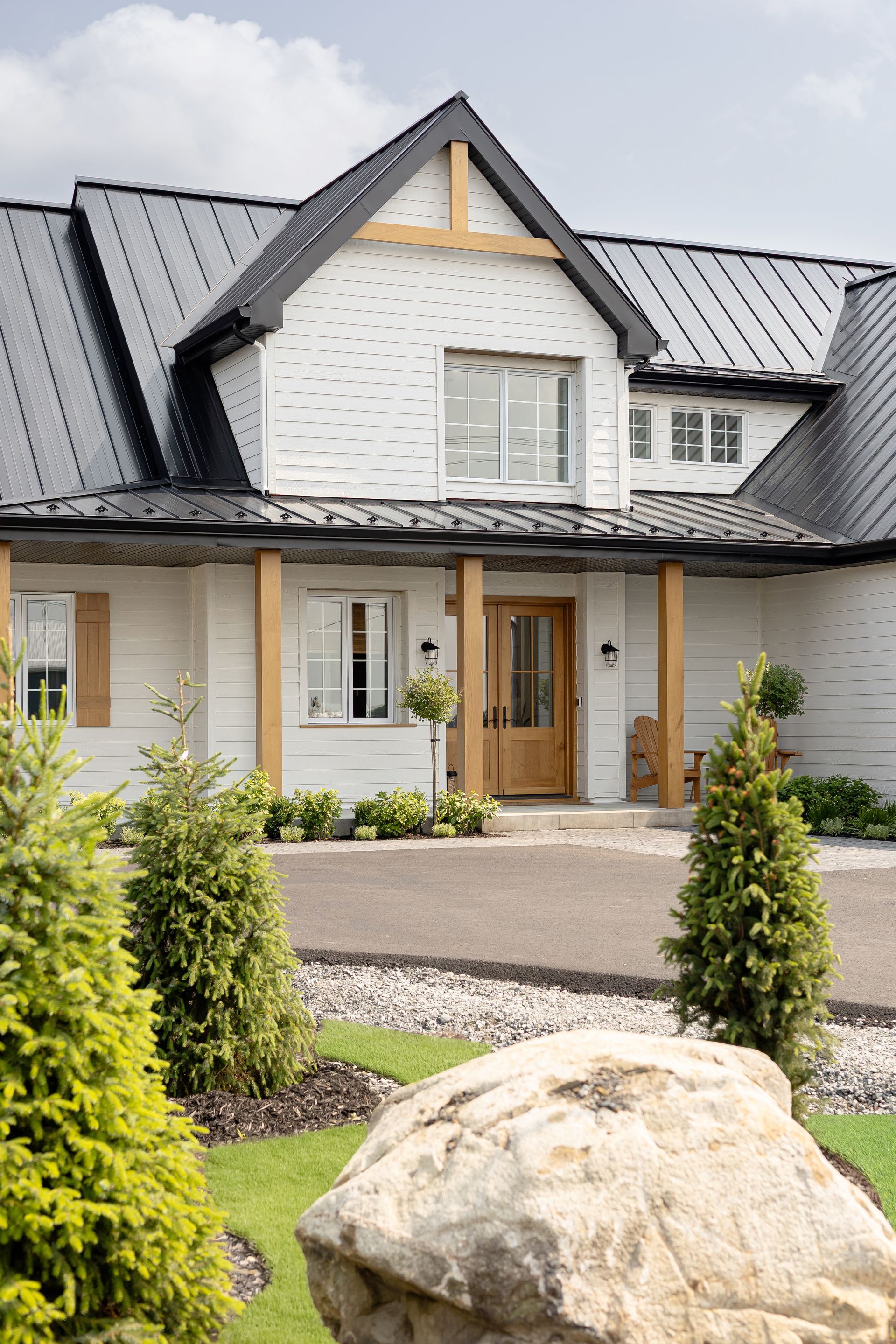 Ferme blanche avec toit noir, accents en bois et aménagement paysager vert.