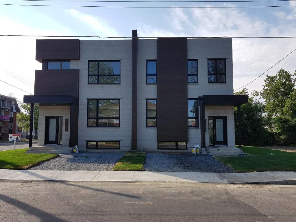 Duplex moderne à deux étages avec des accents brun foncé et des fenêtres à cadre noir, sur un terrain de gravier et d'herbe.