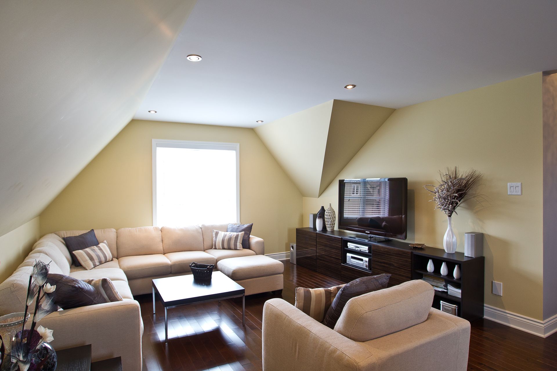Salon confortable avec canapé sectionnel beige, meubles en bois foncé et télévision dans une pièce aux plafonds inclinés.