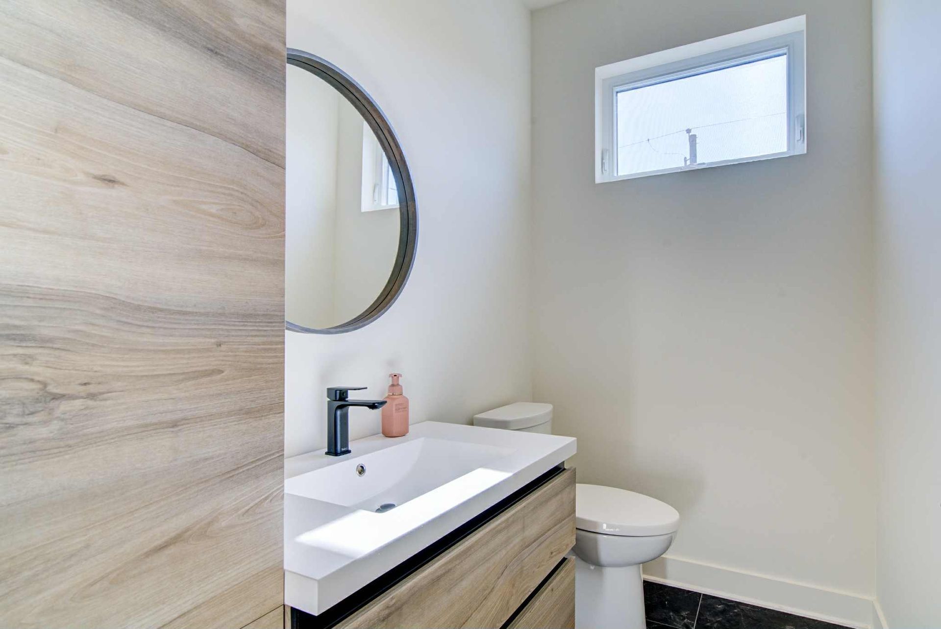 Salle de bain aux murs blancs, avec meuble-lavabo, toilettes et fenêtre. Un miroir rond est suspendu au-dessus du lavabo.