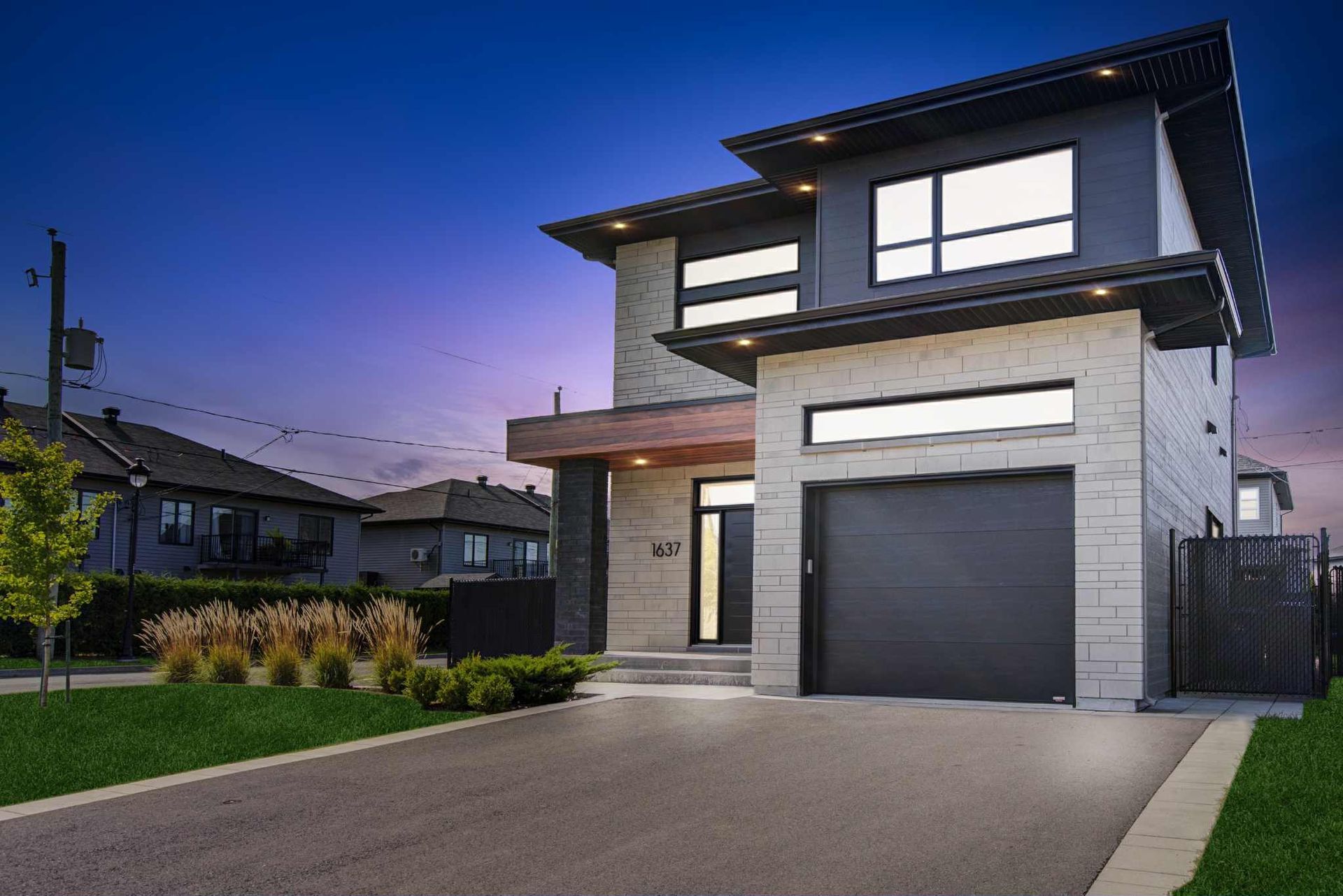 Maison moderne à deux étages avec façade grise, porte de garage noire et allée au crépuscule.