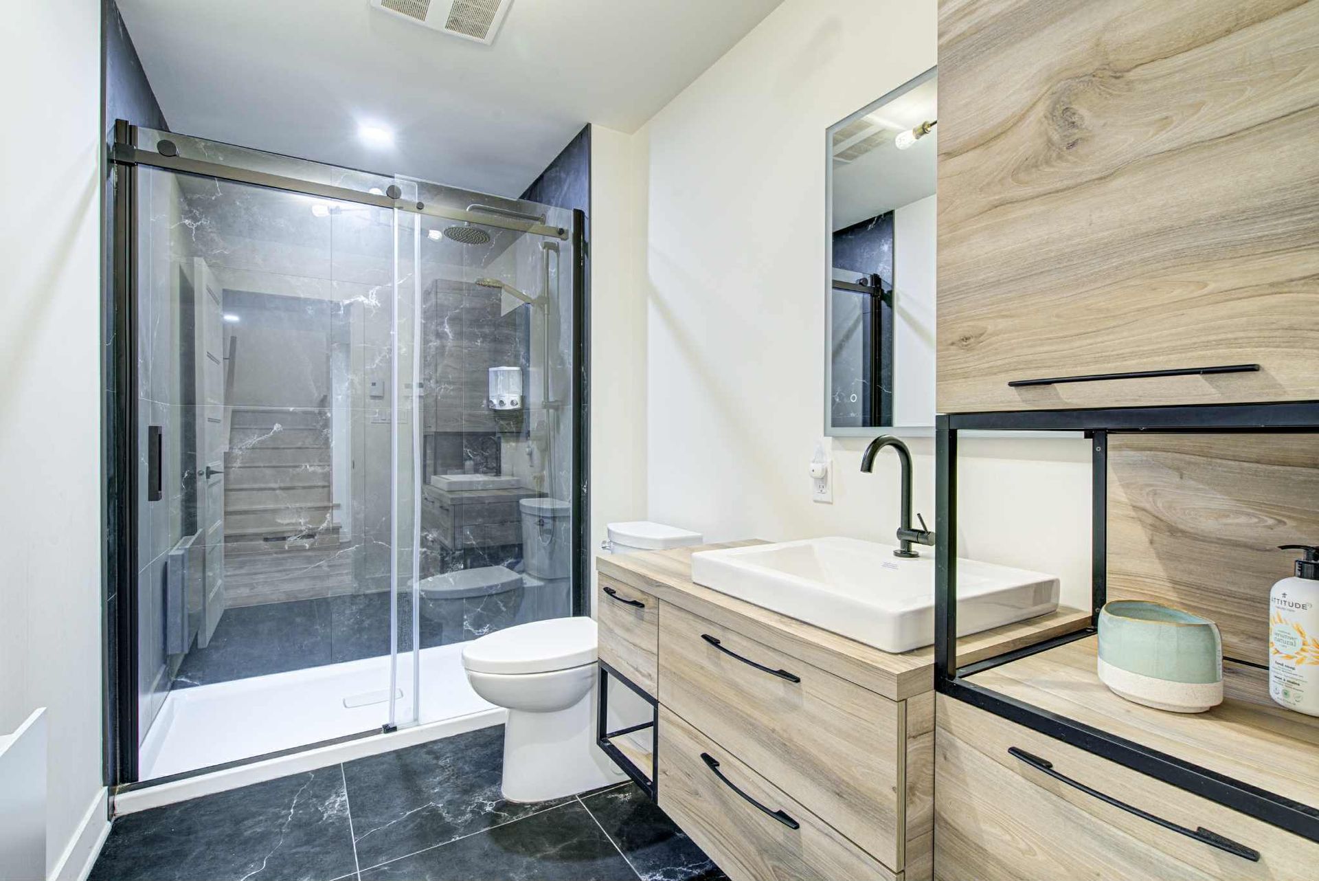 Salle de bain moderne avec douche en verre, vanité en bois et luminaires noirs.