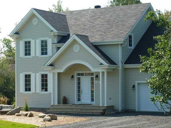 Maison à deux étages avec bardage clair, toit foncé, boiseries blanches et véranda. Allée en gravier, ciel bleu.