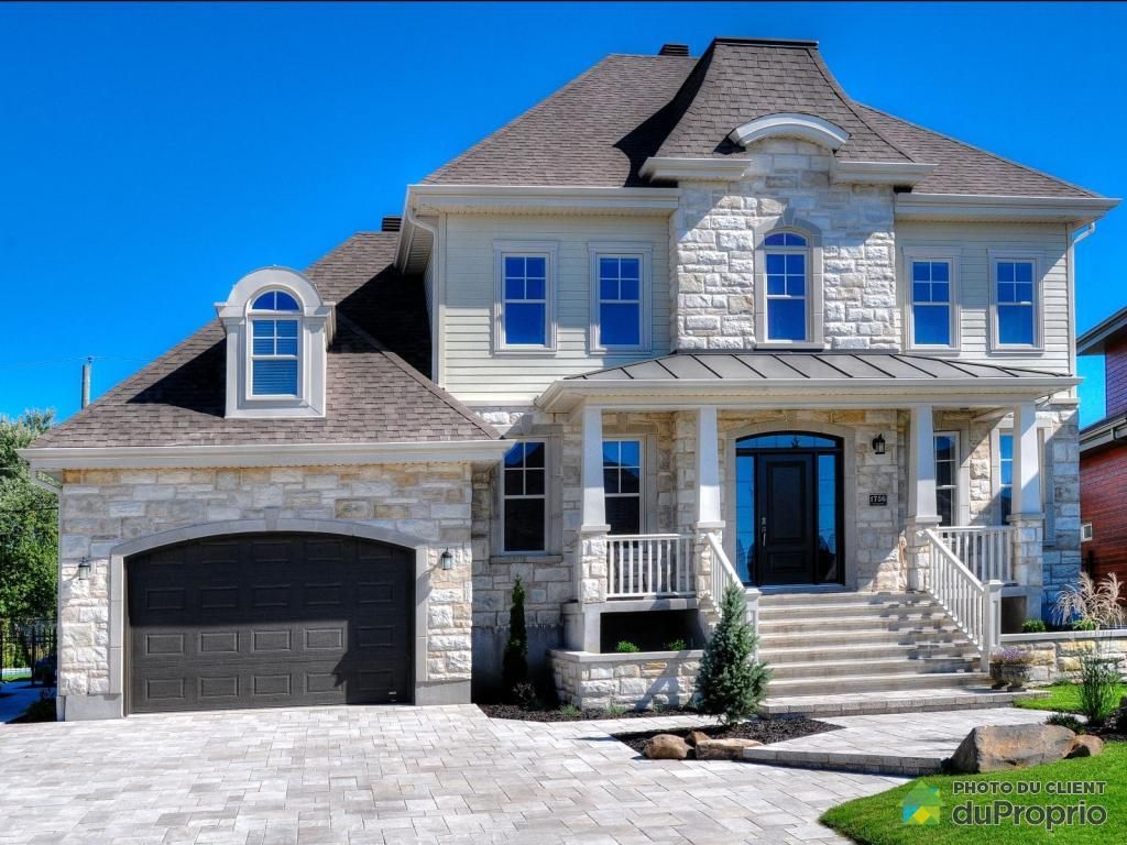 Maison en pierre beige à deux étages avec une porte de garage foncée, des fenêtres bleues et un porche avant.