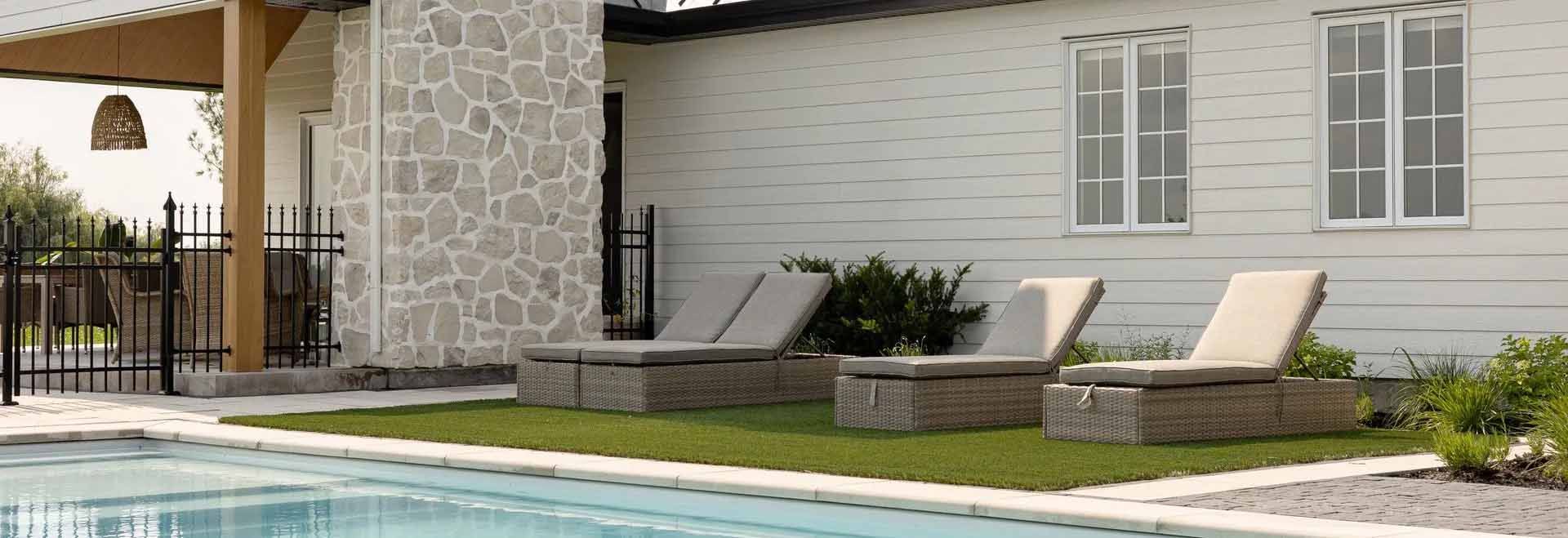 Chaises longues au bord d'une piscine sur une zone herbeuse, à côté d'un bâtiment en briques blanches.