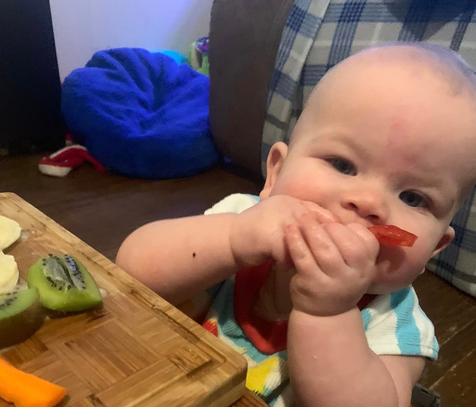 A baby is sitting in a high chair eating fruit from a cutting board.