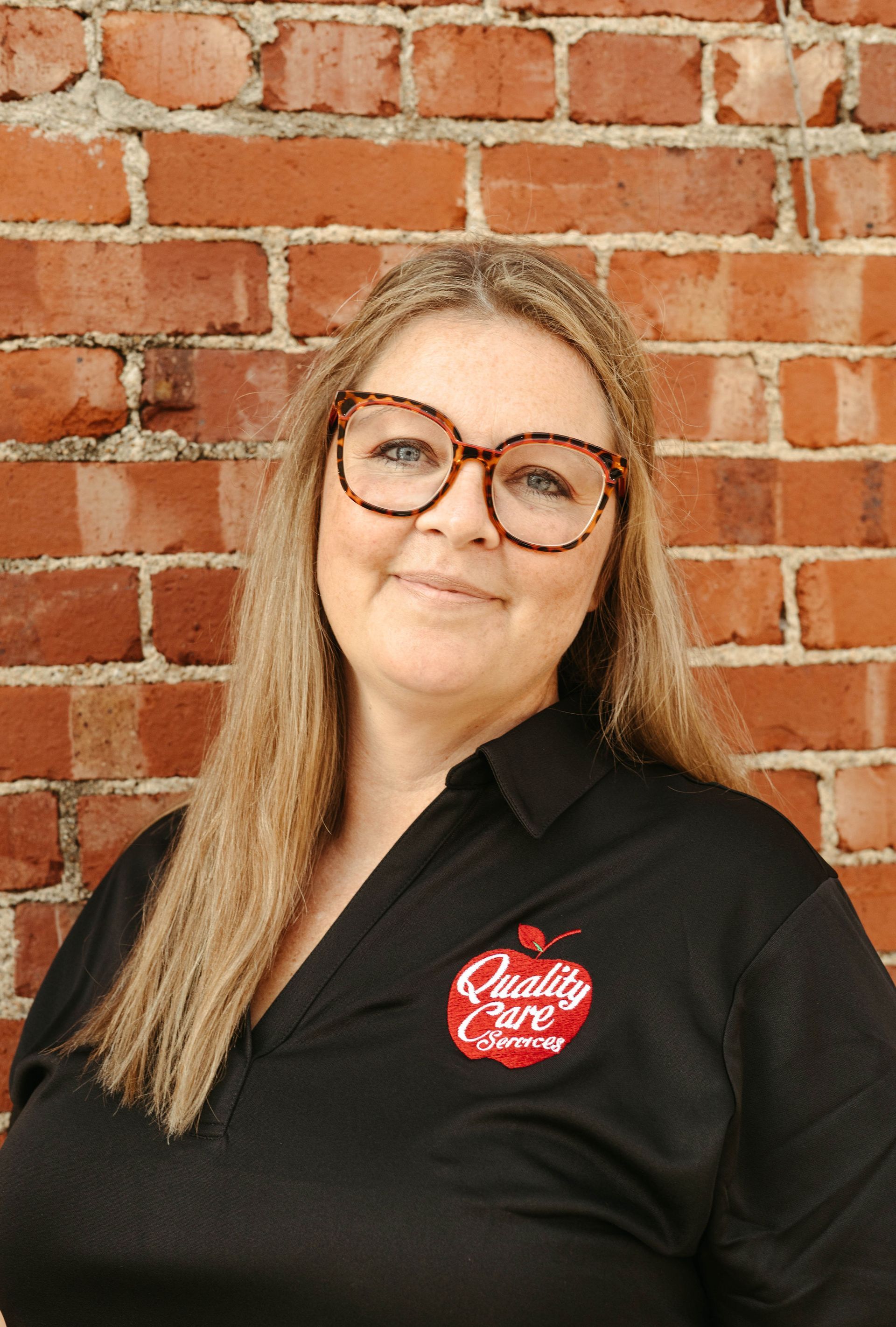 A woman wearing glasses and a black shirt is standing in front of a brick wall.