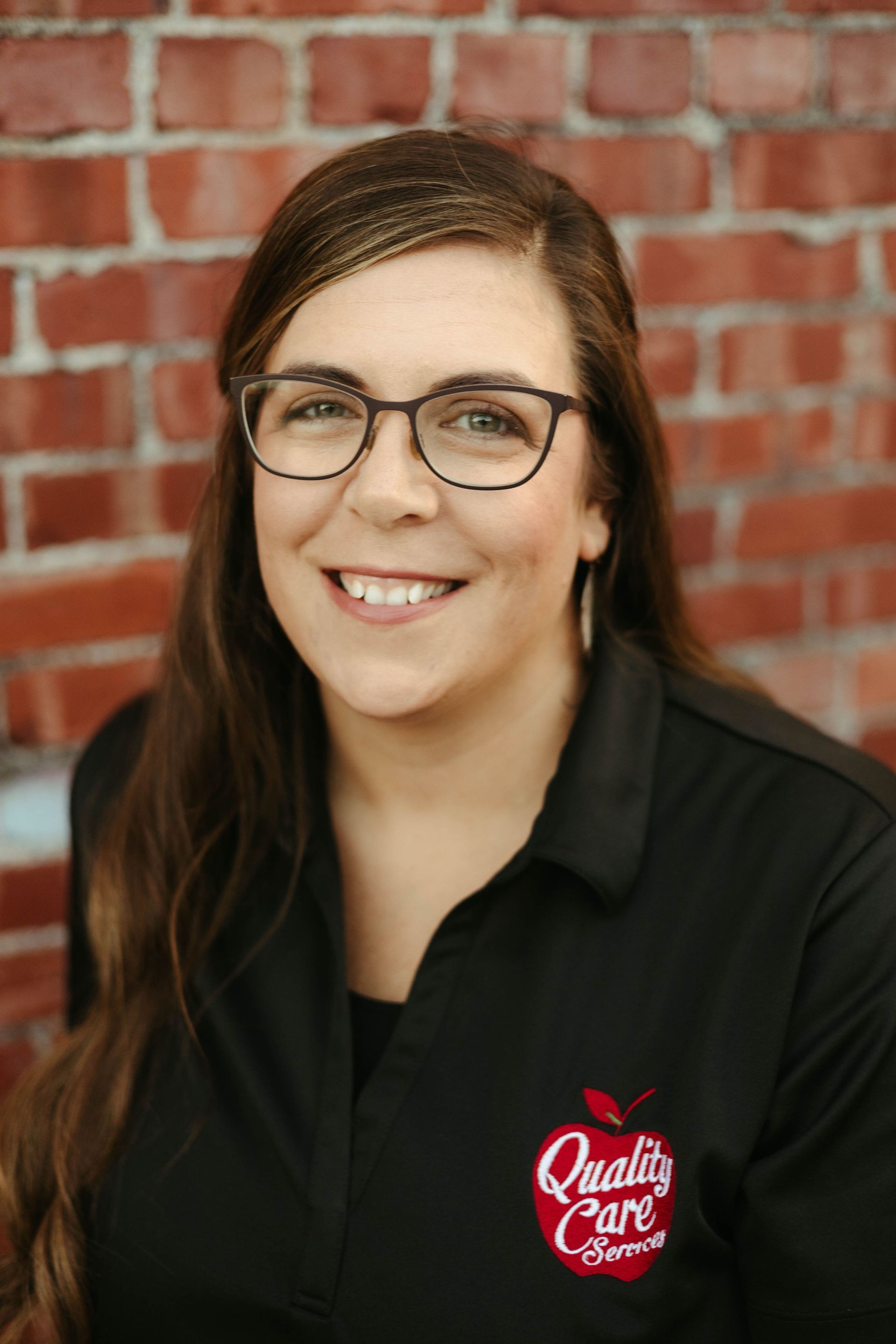 A woman wearing glasses and a black shirt is smiling in front of a brick wall.