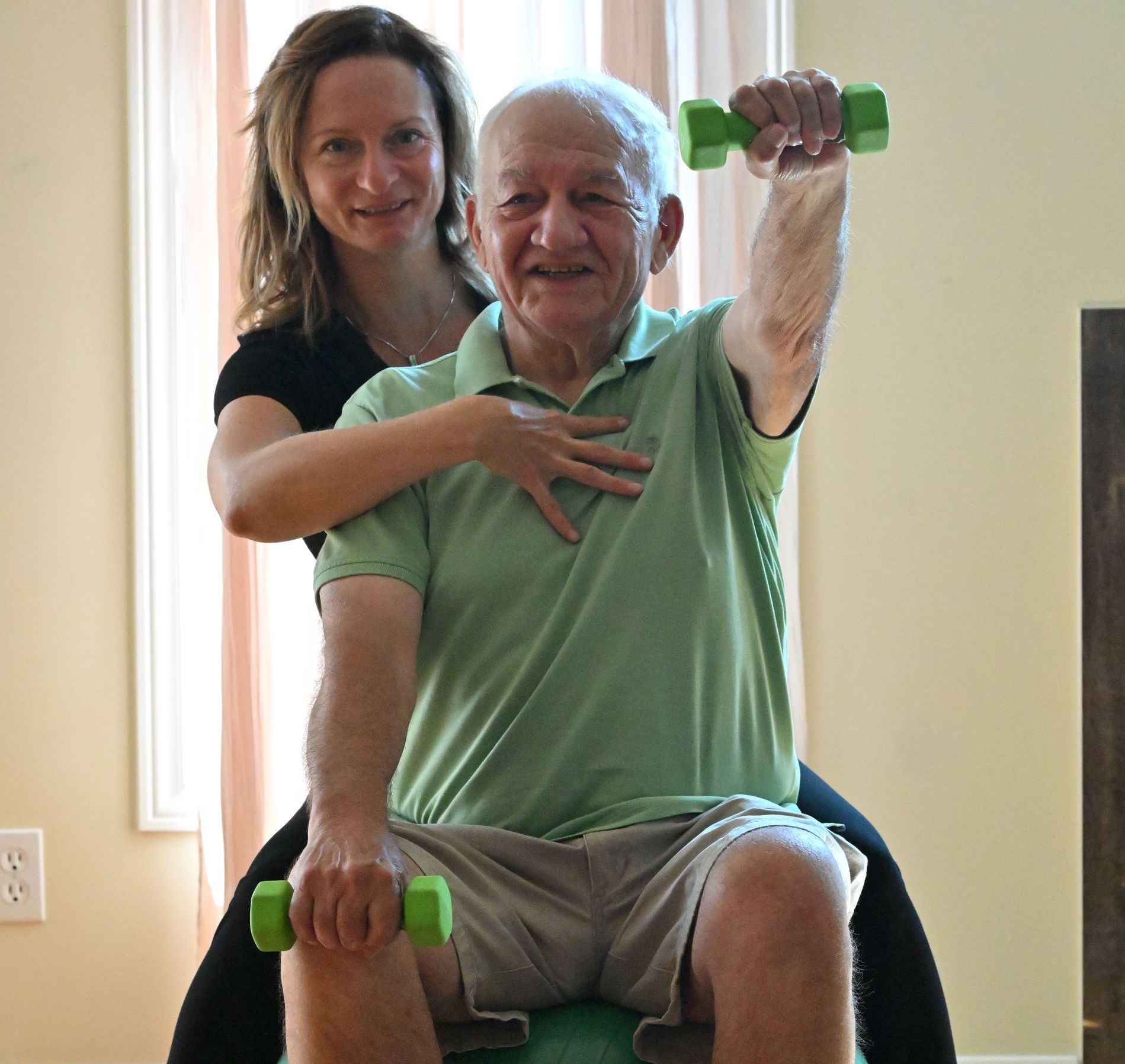 Physiotherapist supporting her elderly client. They are both sitting on a large inflated stability ball. She is sitting behind him and reaching around his right side to gently support him in lifting his chest to elongate his spine while he lifts a weight with left arm extended out in front of him.