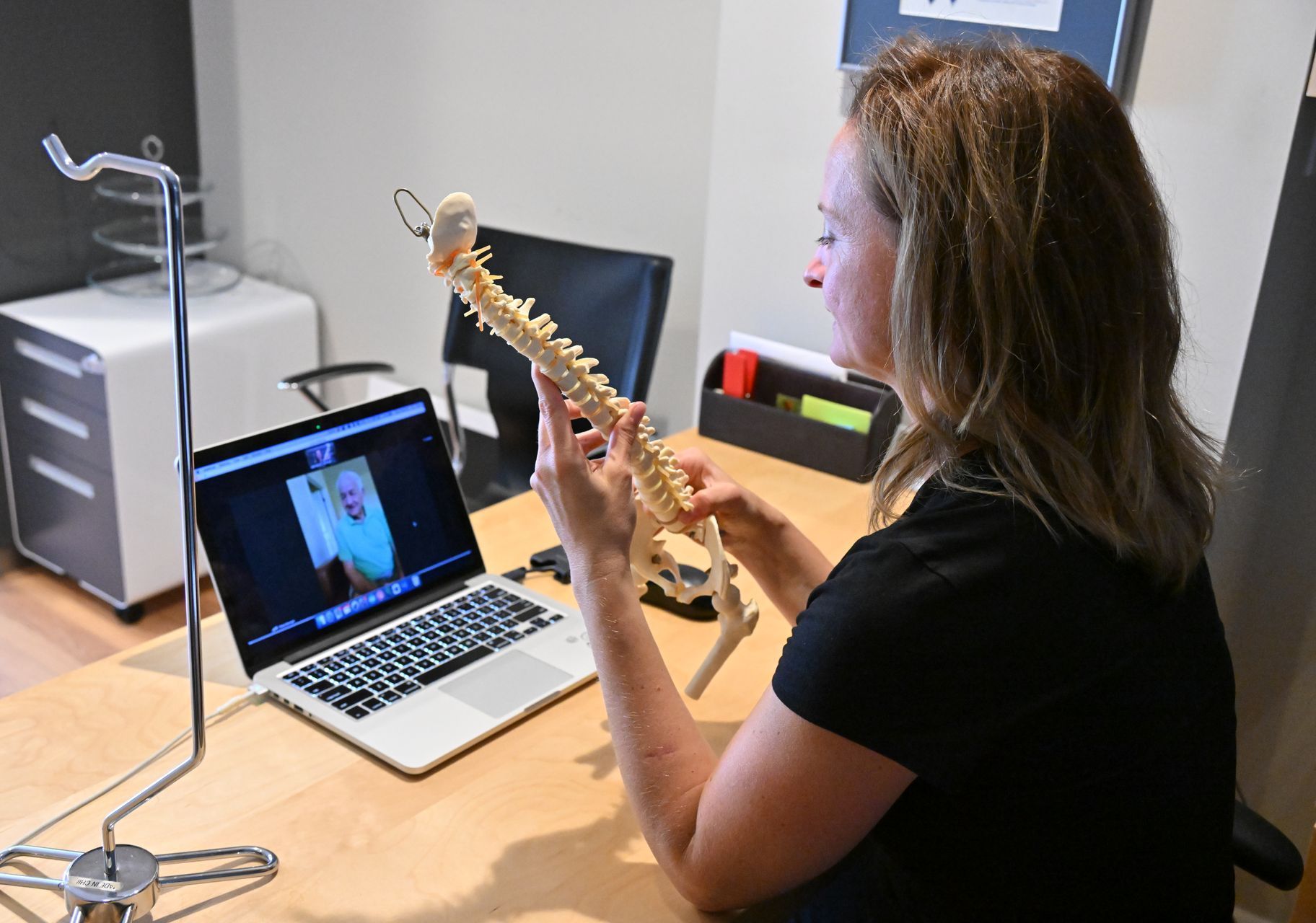 Physiotherapist engaged in a virtual consultation. Her laptop is open in front of her with her client on screen. She holds a model of a spine as she explains something to her client.