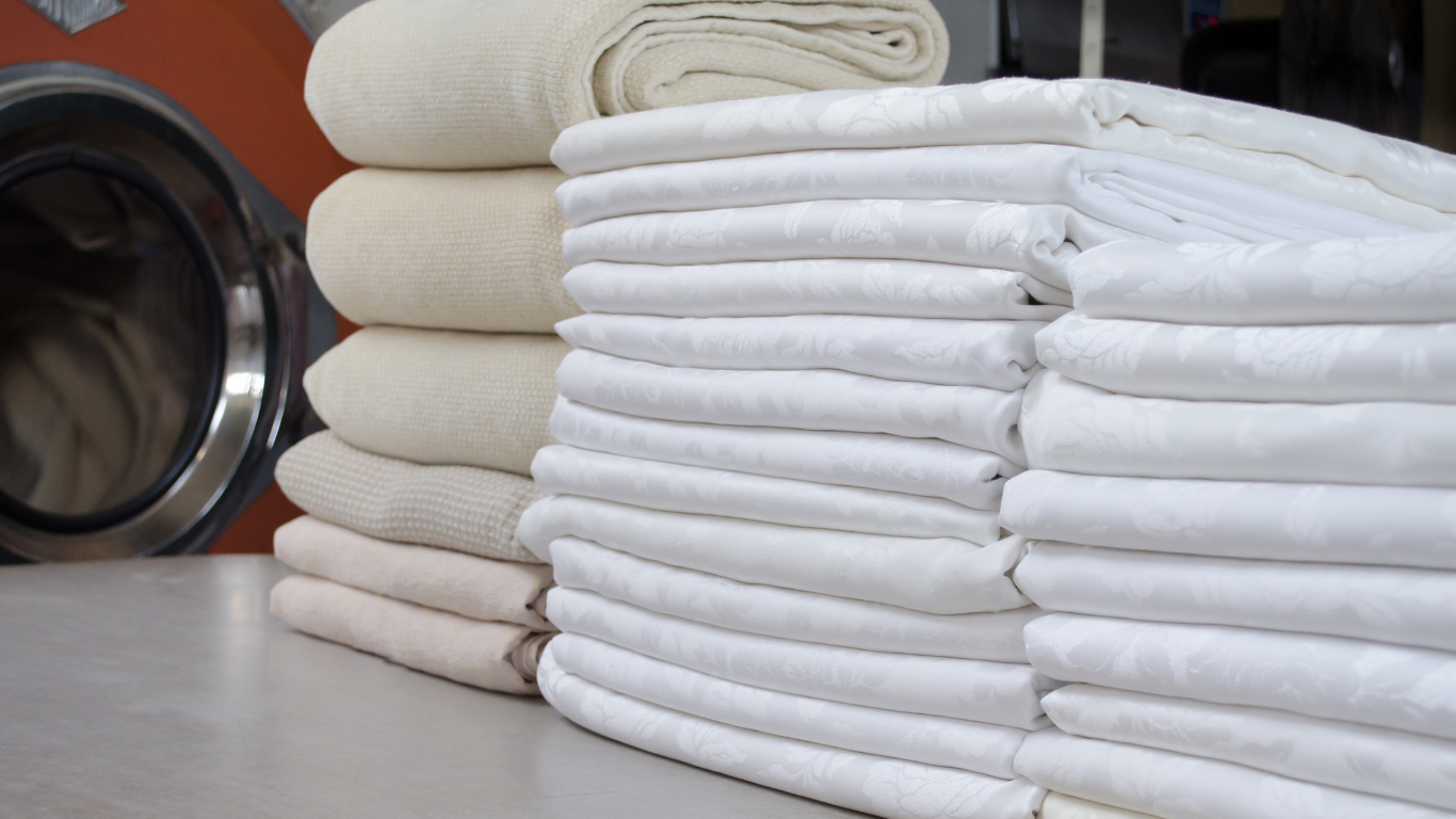 Stacks of neatly folded white and cream-colored linens sit on a table next to a commercial laundry machine.
