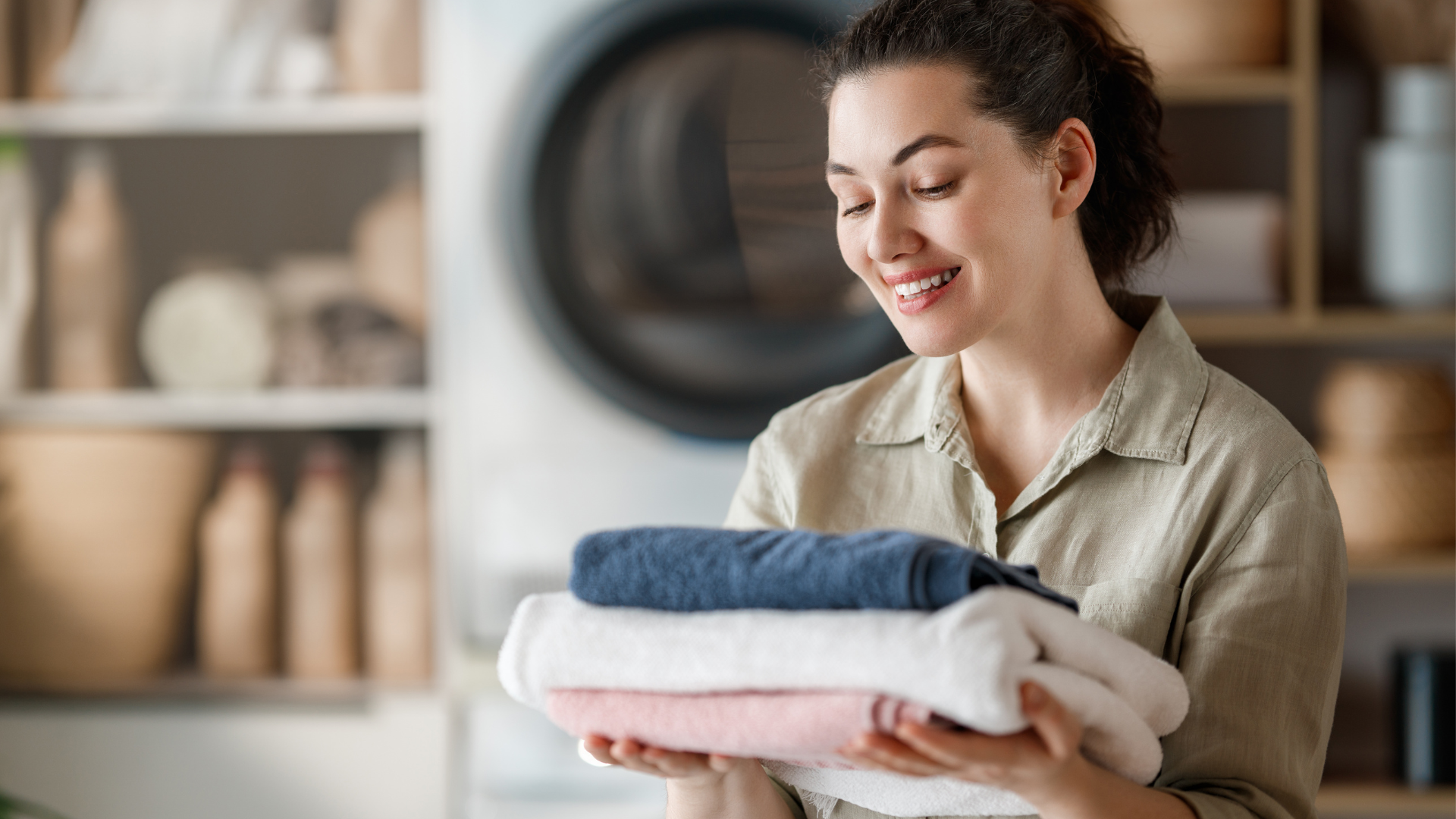 A person smiles while holding a neat stack of folded blue, white, and pink towels in a laundry room.