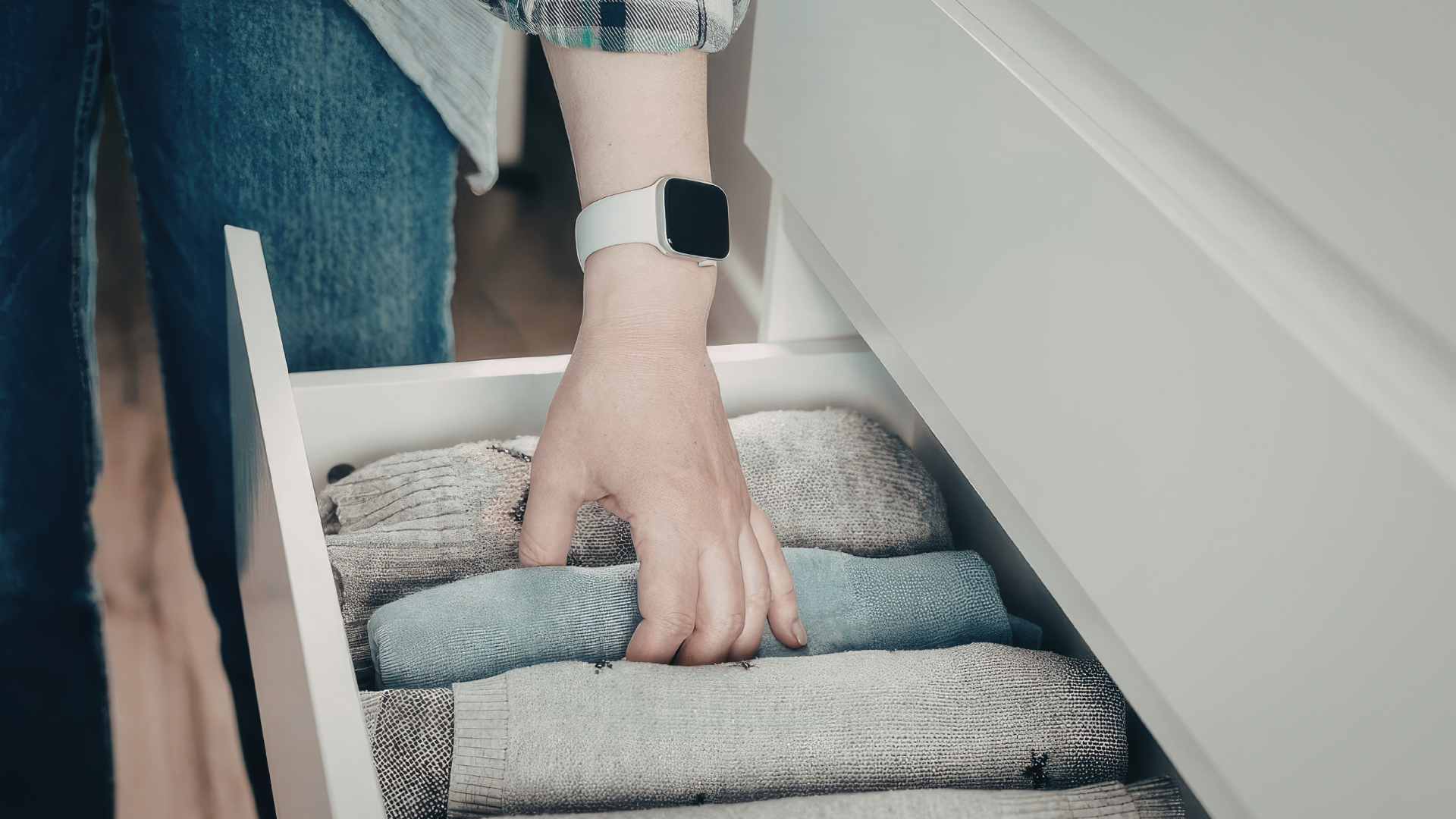 A person wearing a white smart watch reaches into a white drawer to organize folded gray and blue textiles.