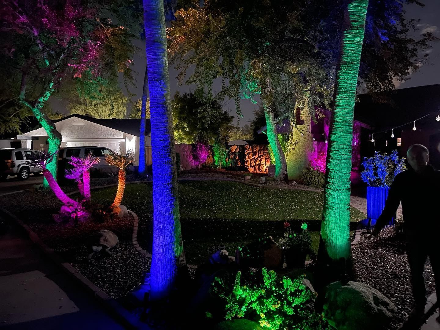 A person is standing in front of a house with trees lit up with colored lights.