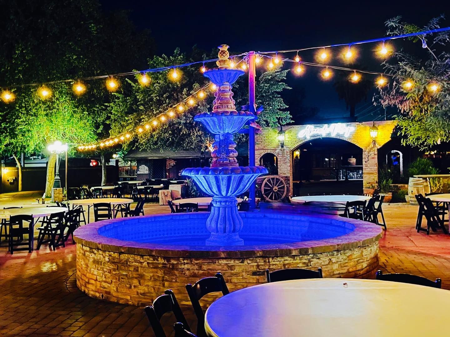 A fountain is lit up at night with tables and chairs around it.