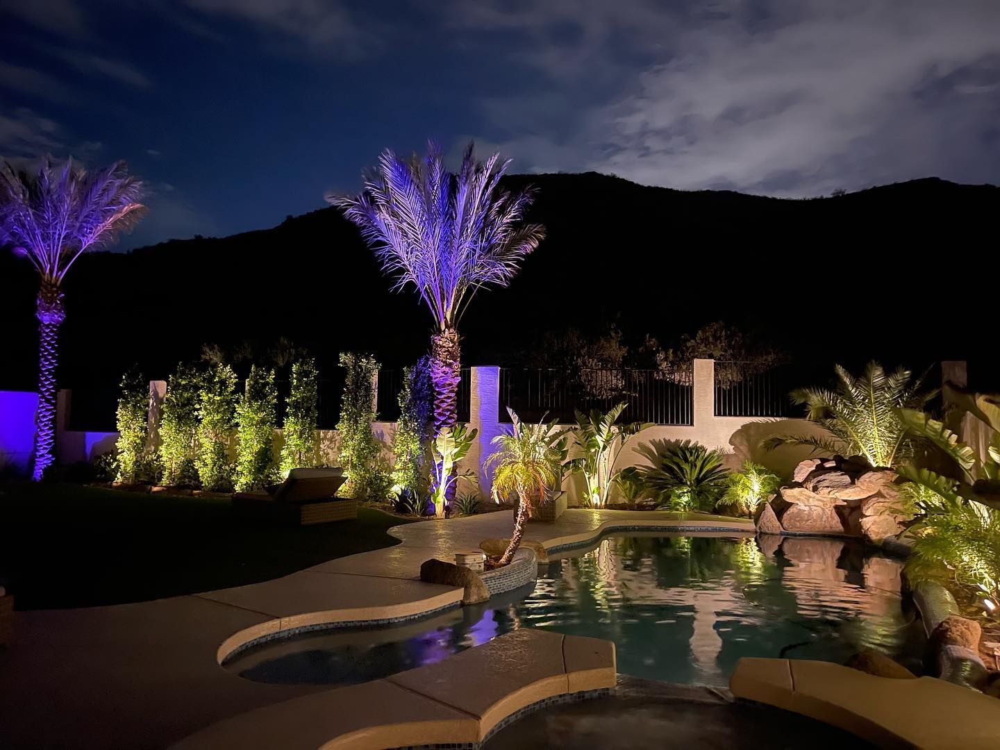 A swimming pool is lit up at night with palm trees and mountains in the background.