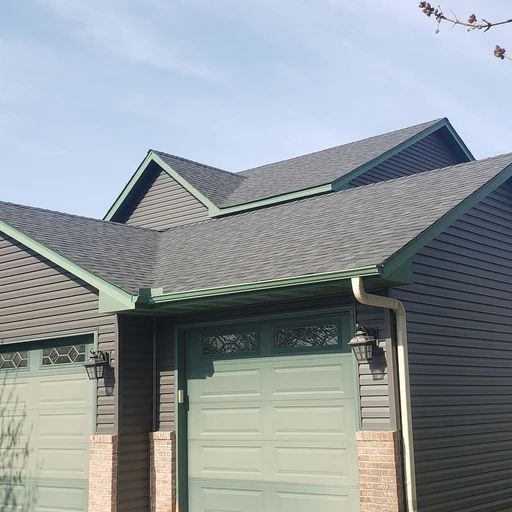 Gray house with green trim, garage doors, and roof against a blue sky.