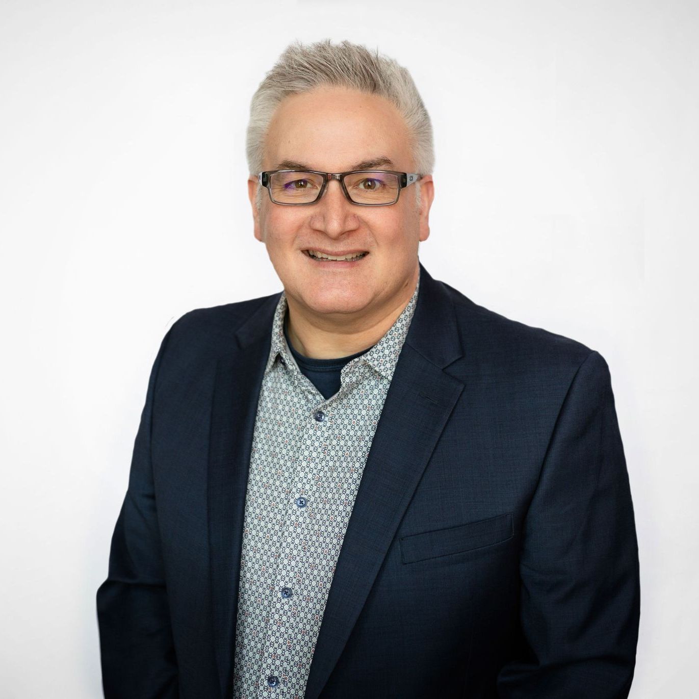 Man in glasses and suit jacket, smiling, standing against a white backdrop.