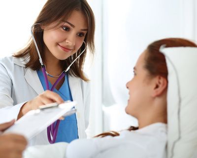 Doctor using a stethoscope on a patient in a hospital bed, smiling, holding a clipboard.