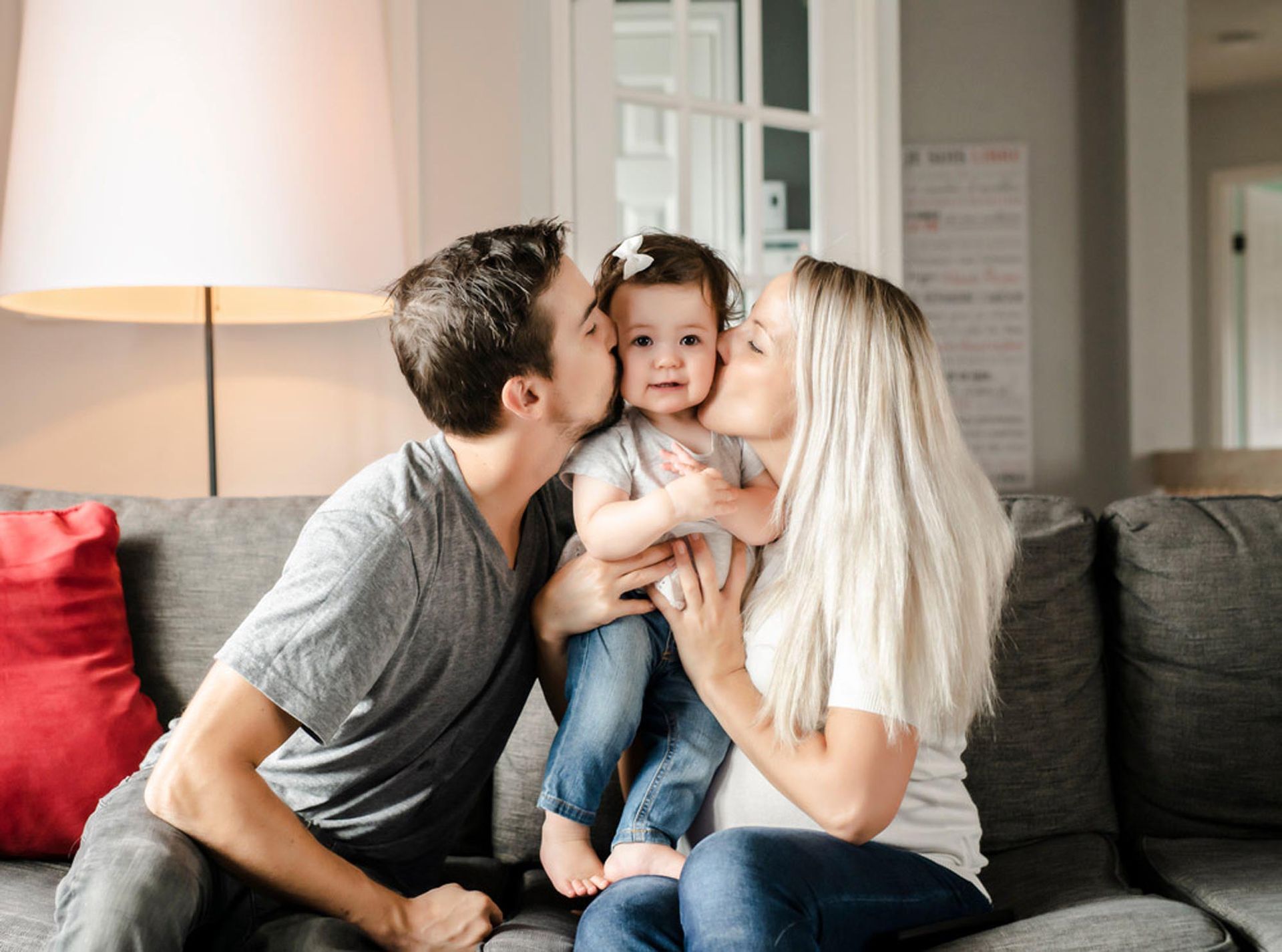 Parents kissing a baby on cheeks while sitting on a couch. The baby wears a headband.