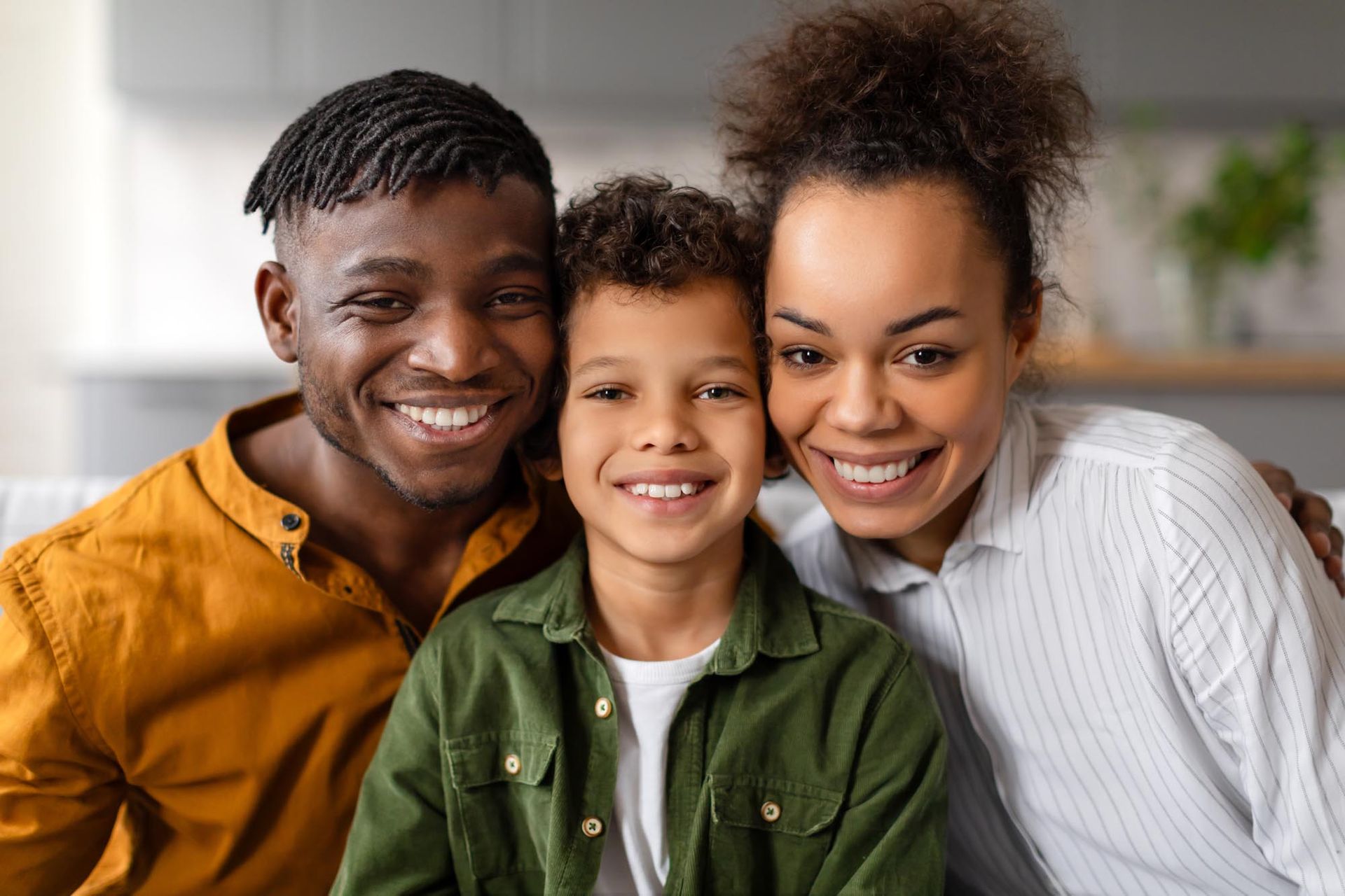 Family smiling and posing together. Man in orange, boy in green, woman in white, indoor setting.