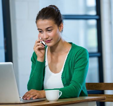 Woman in green cardigan on phone, working on laptop, with coffee cup.