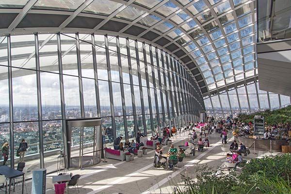 Interior of a glass building with people, plants, and a city view.