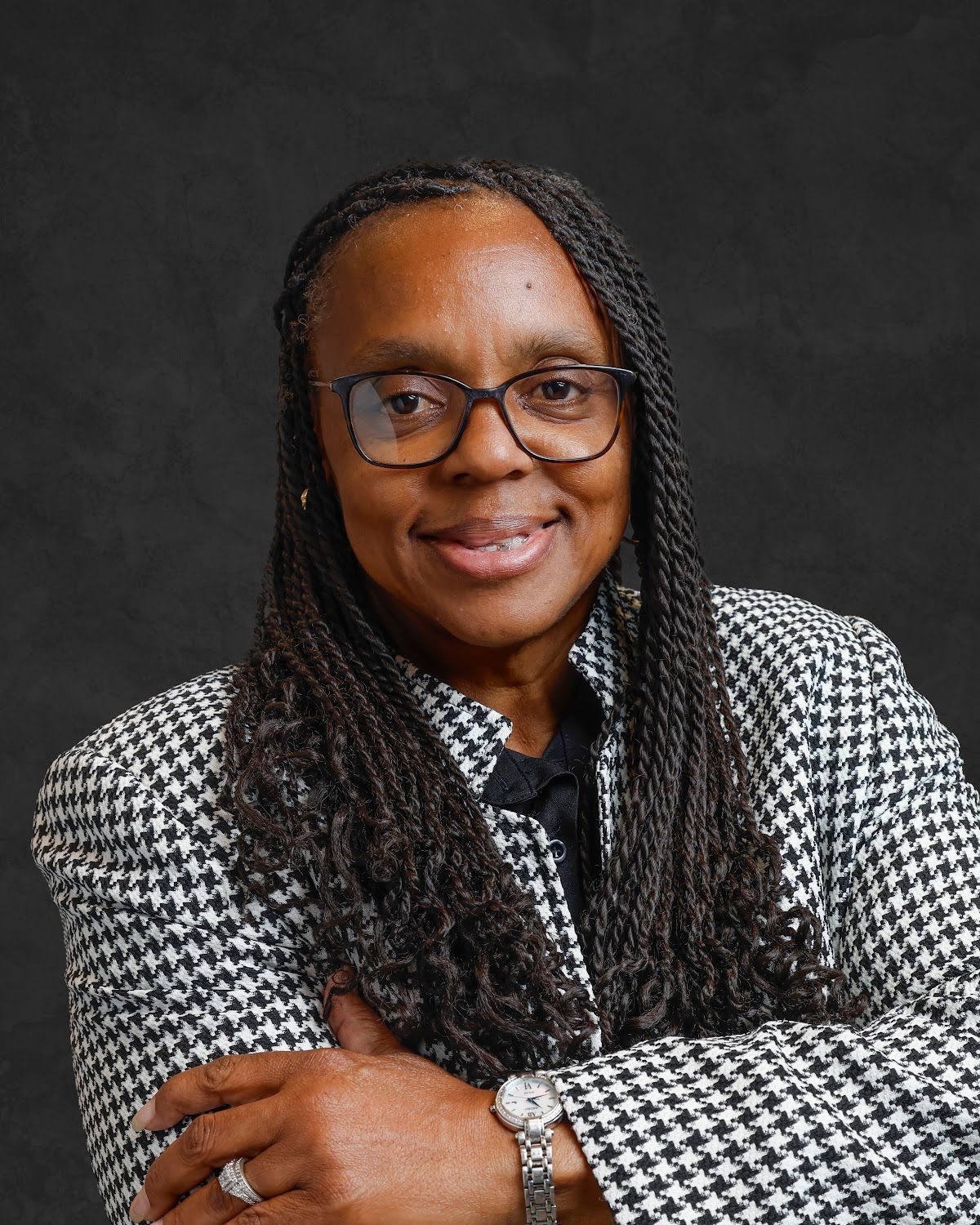Woman with glasses, braids, wearing a black and white checked blazer, arms crossed, smiling.