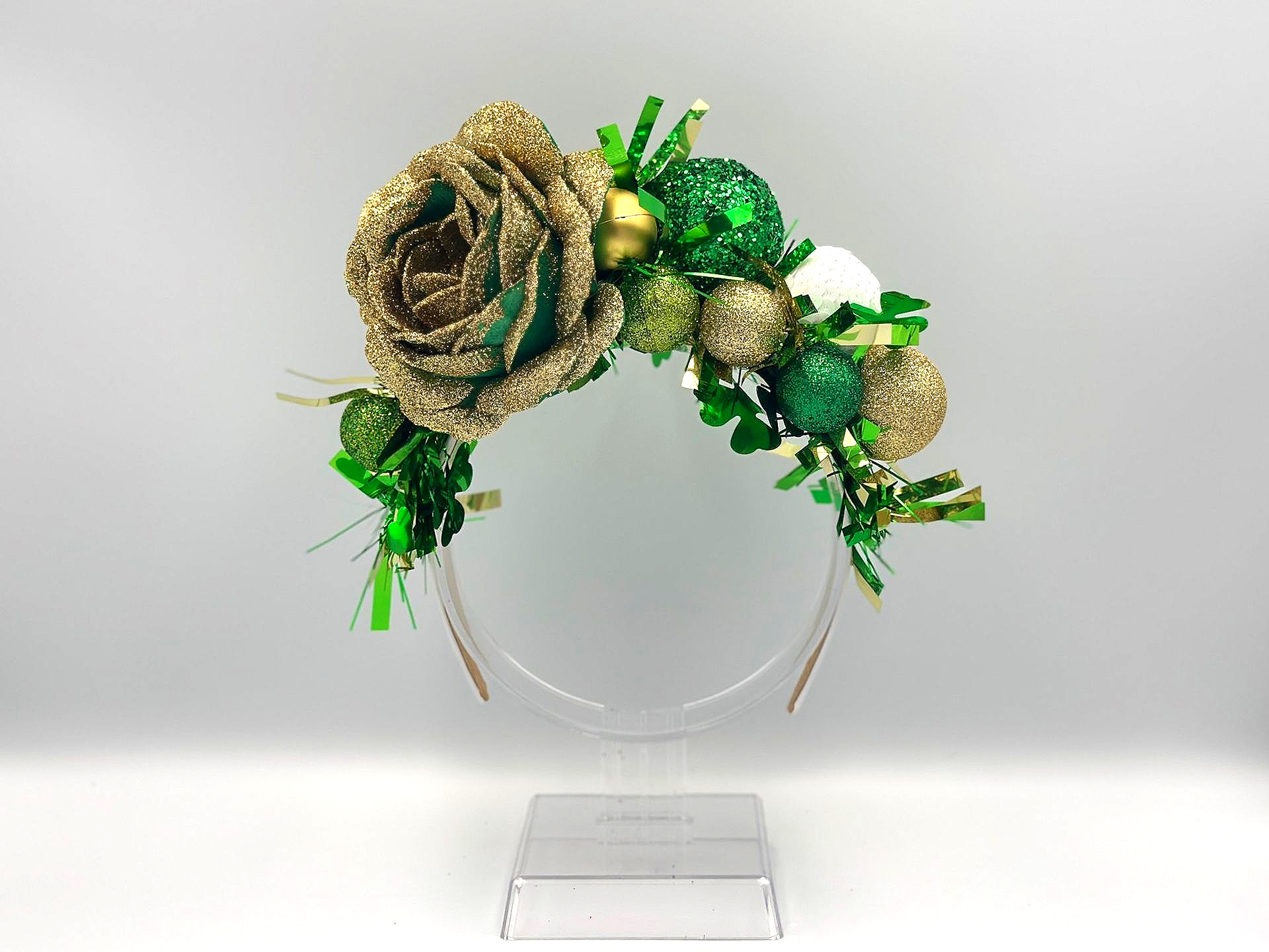 Green and gold floral headband on a clear display stand. Includes glittery balls and a large rose.