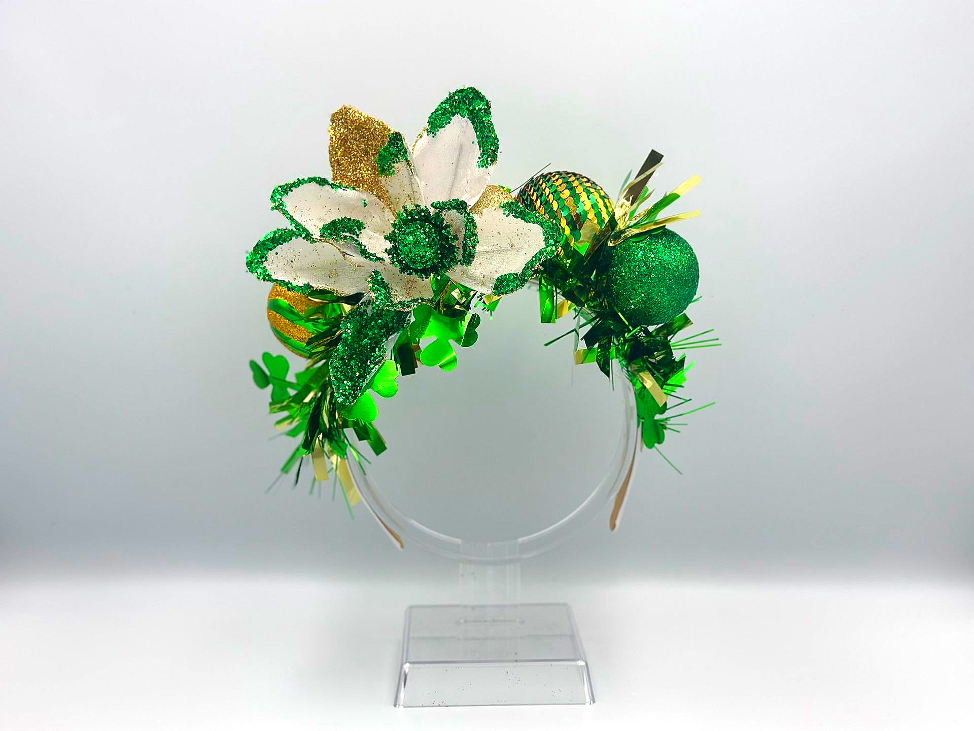 Green and gold sparkly St. Patrick's Day headband with flowers and ornaments, on a clear stand.