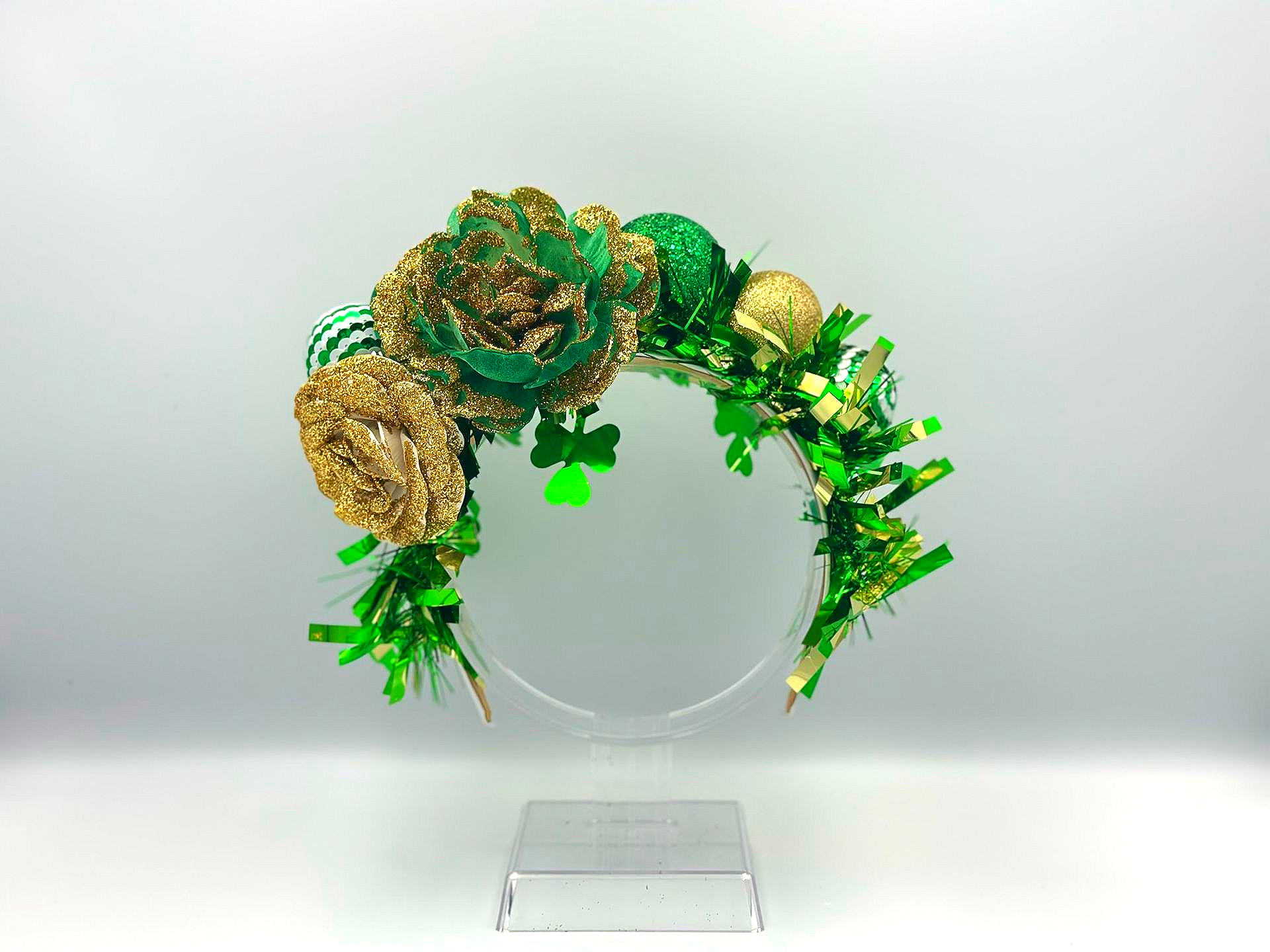 St. Patrick's Day headband with green and gold decorations, including shamrocks, and flowers on a clear plastic stand.