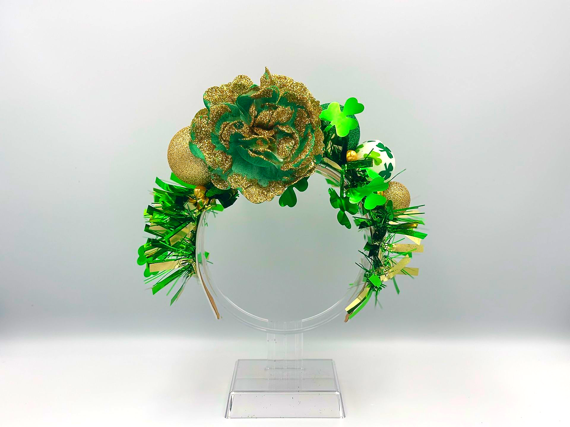Green and gold St. Patrick's Day headband with large glittery flower, shamrocks, and tinsel on clear stand.