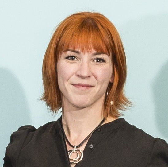 A woman with red hair is smiling while wearing a black shirt with a silver necklace