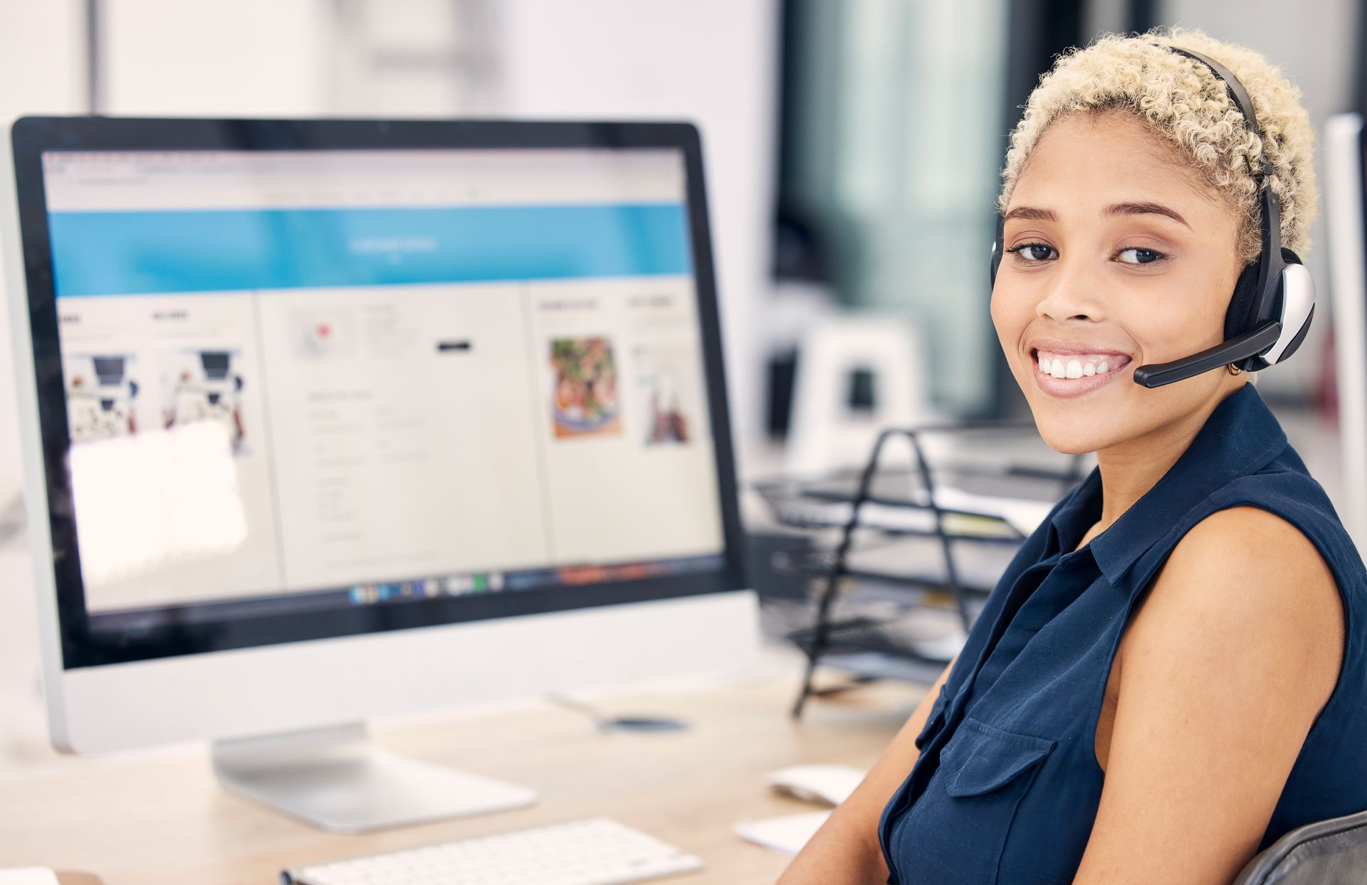Woman wearing a headset, smiling at a computer. She is in an office, working.