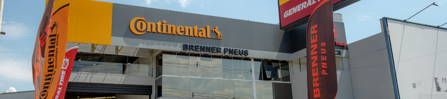 Mecânico de macacão amarelo empurrando um pneu em direção a um SUV cinza em uma garagem.