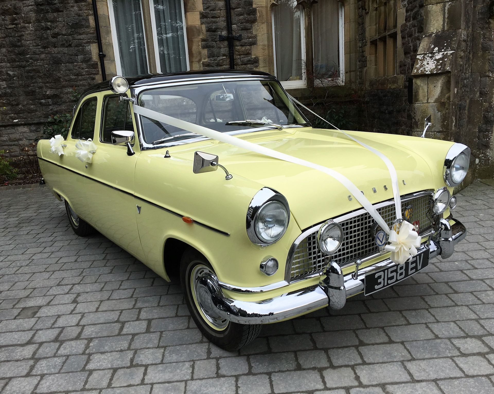 Close-up of a 1959 Ford Consul in its original primrose yellow, decorated with white bows, ready to pick up the newlyweds