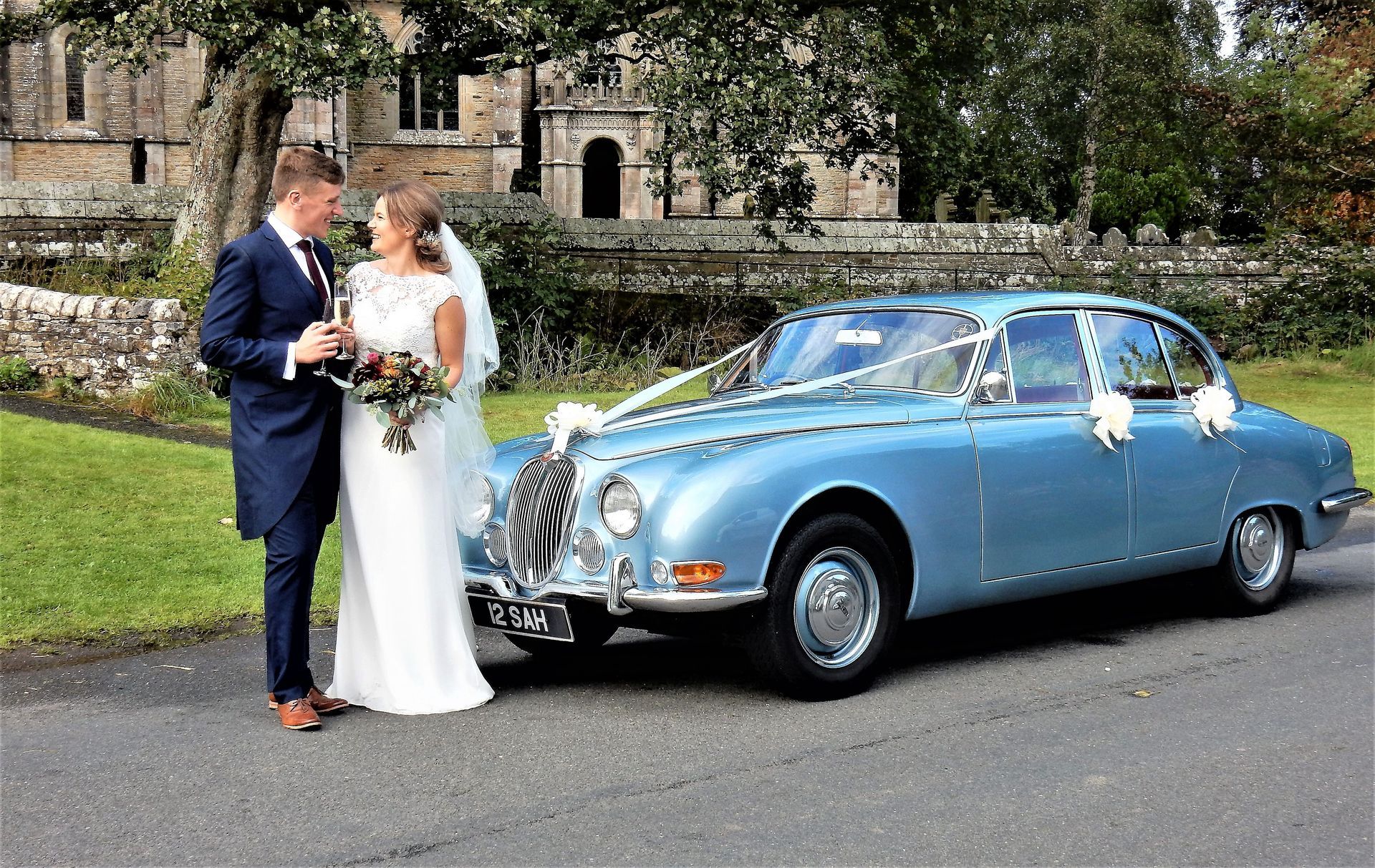 Newlywed couple joyfully toasting with a glass of fizz, gazing at each other beside their adorned light blue Jaguar MK2 wedding car with white bows.
