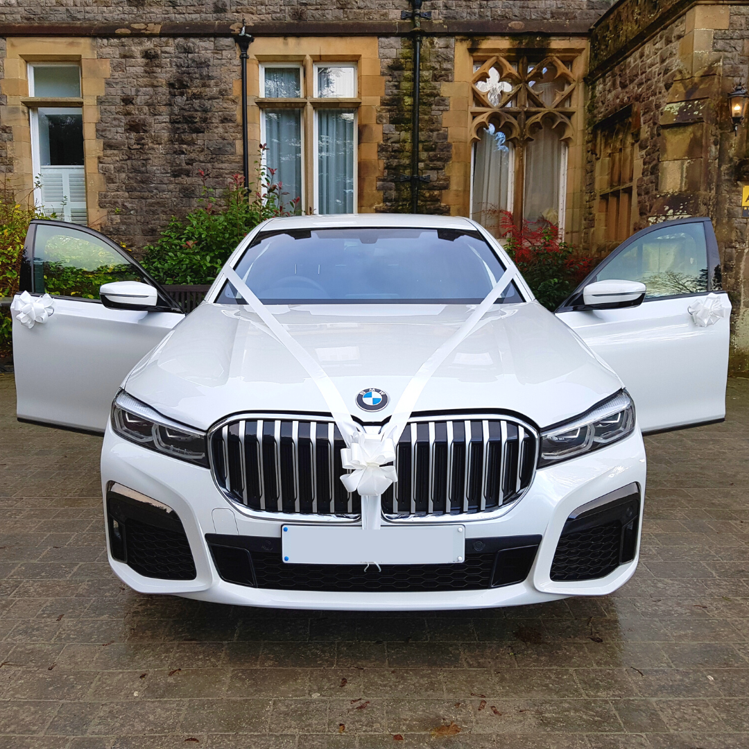 Close-up of a pristine white BMW 7 Series adorned with white bows, doors wide open, and additional bows on display.