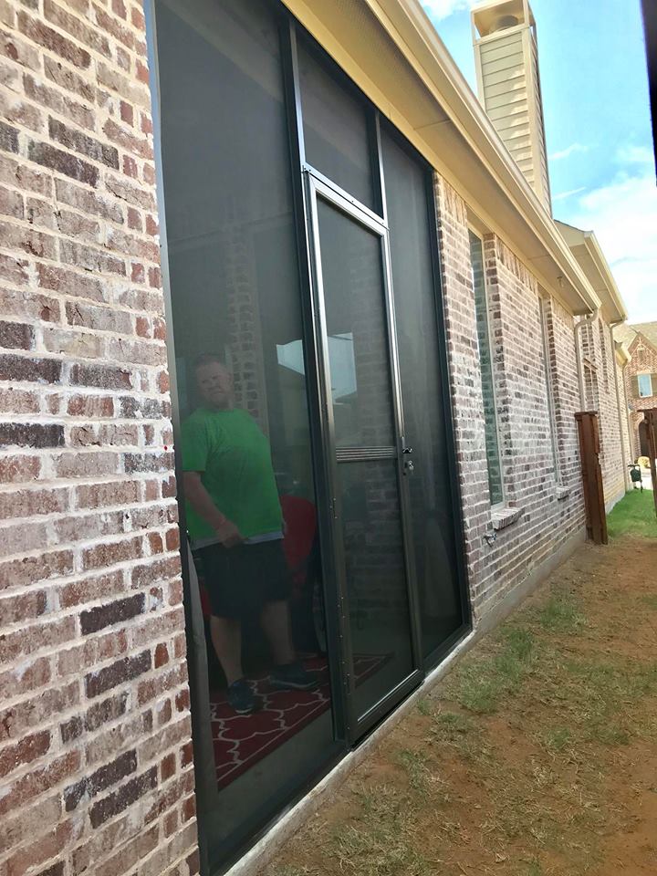 A man in a green shirt is standing in front of a screen door.