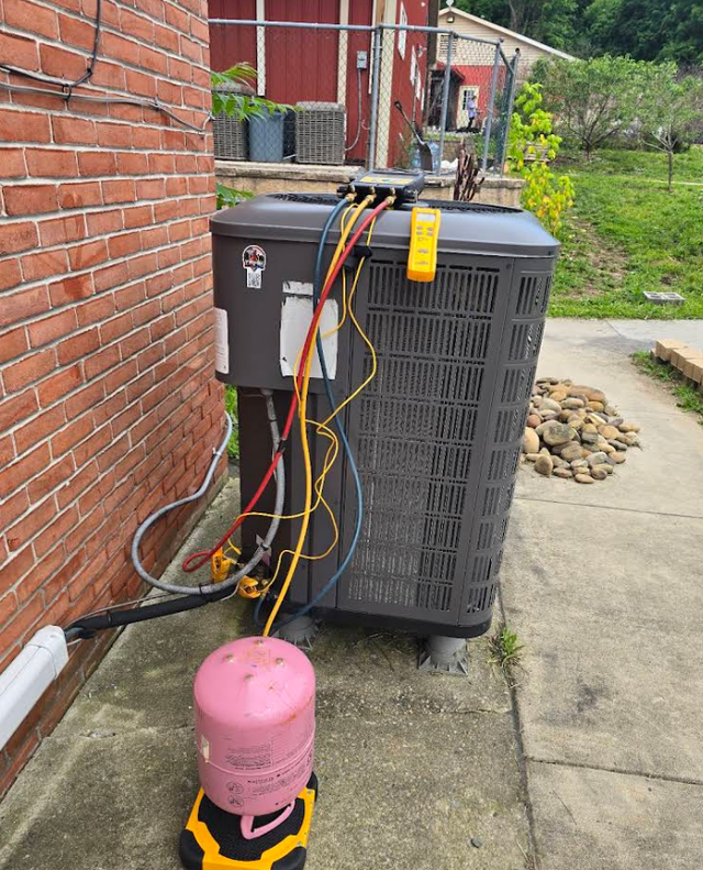 Outdoor HVAC unit beside a brick wall with attached service hoses and a pink refrigerant tank in front