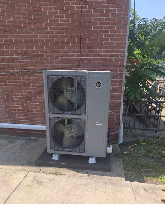 Outdoor HVAC unit beside a brick wall on a concrete pad, with two fans visible.